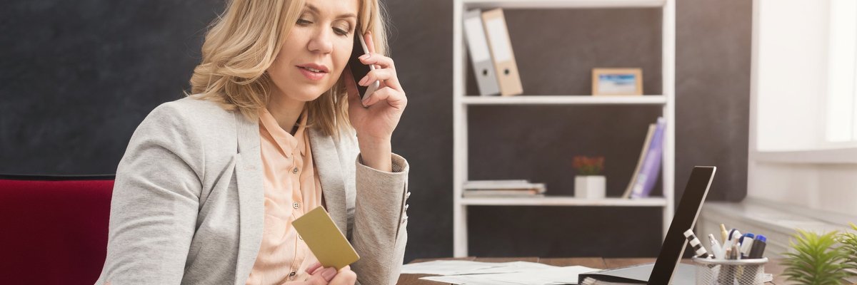 Woman in business suit sitting in front of laptop looking at a credit card and talking on the phone.