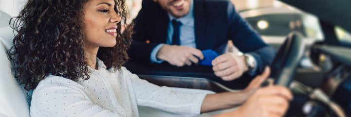A woman sitting in the driver's seat of a new car and talking to a salesman through the window.