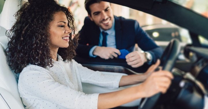 A woman sitting in the driver's seat of a new car and talking to a salesman through the window.