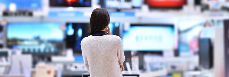 A woman standing in an electronics store facing a wall of TVs.