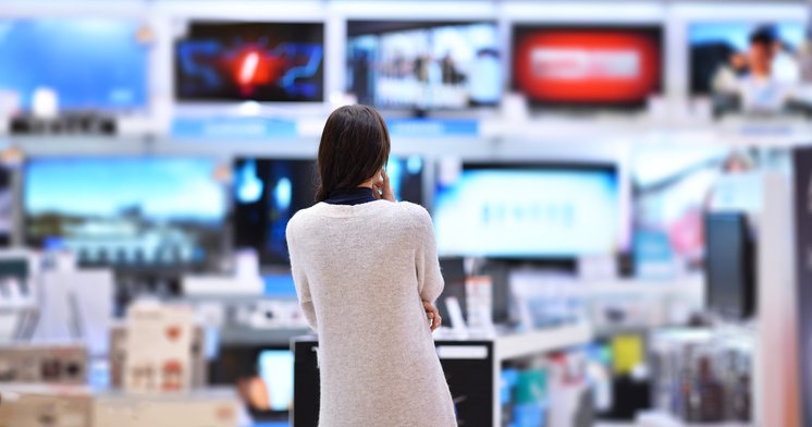 A woman standing in an electronics store facing a wall of TVs.
