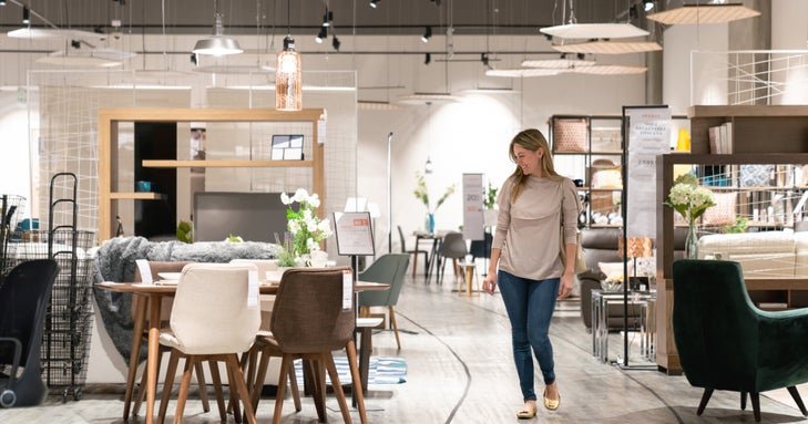 A woman walking around a furniture store and looking at a dining table set.