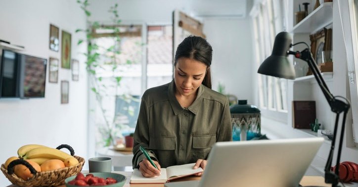 A woman sitting at her kitchen counter next to bowls of fruit and a laptop and writing in a notebook.