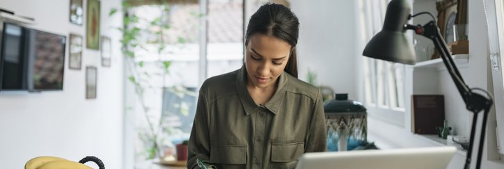 A woman sitting at her kitchen counter next to bowls of fruit and a laptop and writing in a notebook.