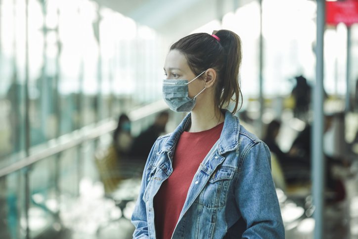 A young woman wearing a medical mask and standing outside an office while looking thoughtfully into space.