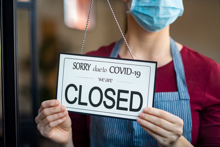 A woman wearing a medical mask and an apron turning a window sign that says the business is closed due to COVID-19.