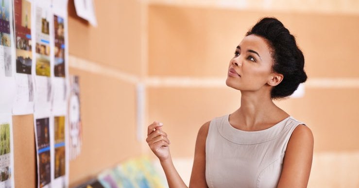 A young businesswoman looking thoughtfully at a wall of pictures in an office.