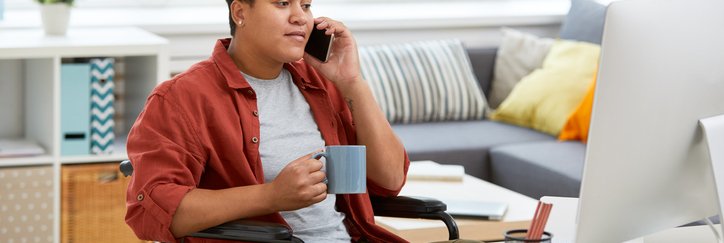 A person sitting in a wheelchair at a home office desk and making a phone call while looking at a computer.