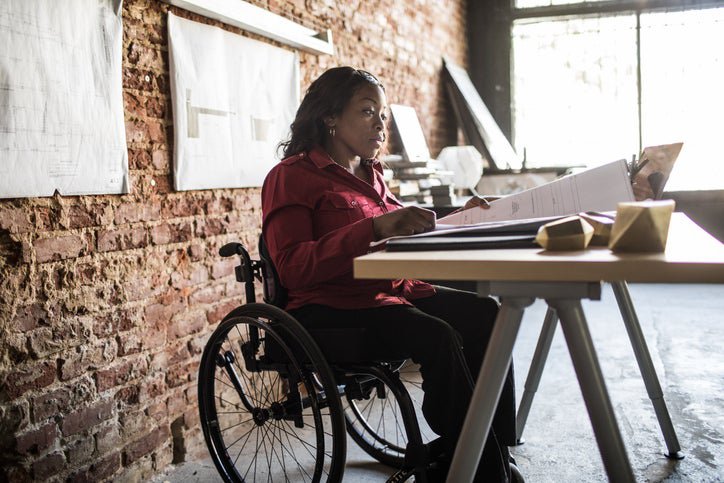 A woman in a wheelchair looking through large documents on a table with more hanging on the brick wall behind her.