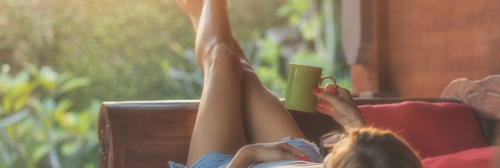 A woman lying down on a patio chair, looking at her sunny backyard, and holding a coffee mug.