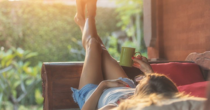 A woman lying down on a patio chair, looking at her sunny backyard, and holding a coffee mug.
