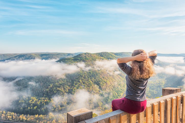 A woman sitting on a railing at a lookout point above forested hillsides wrapped in clouds.