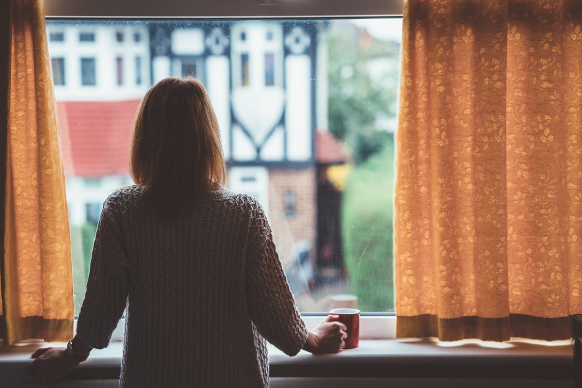 A woman standing at the window in her home and looking out at the neighborhood.