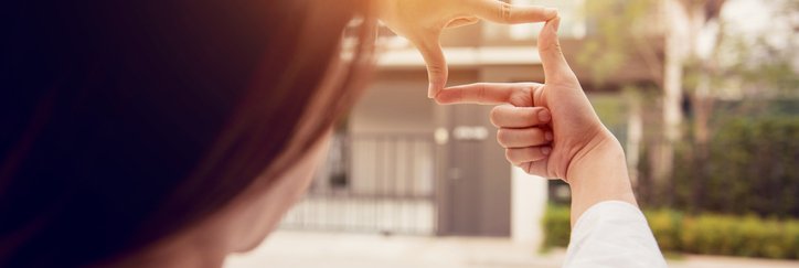 A woman holding her hands up in a square and looking through them at a house on a sunny street.