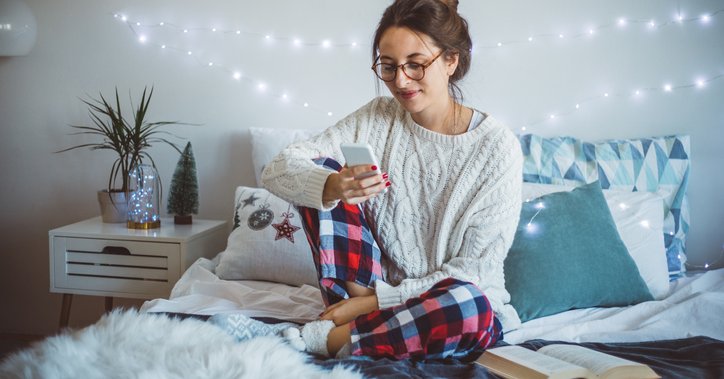 A woman wearing pajamas and sitting on her bed and making a phone call with holiday twinkle lights on the wall behind her.