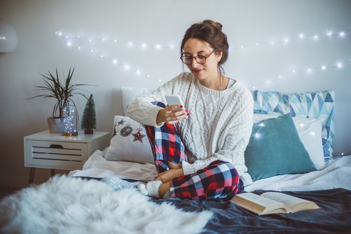 A woman wearing pajamas and sitting on her bed and making a phone call with holiday twinkle lights on the wall behind her.