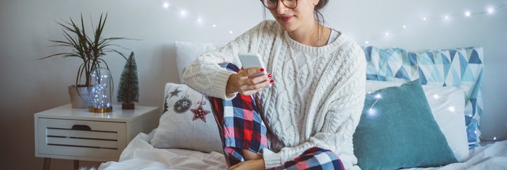 A woman wearing pajamas and sitting on her bed and making a phone call with holiday twinkle lights on the wall behind her.