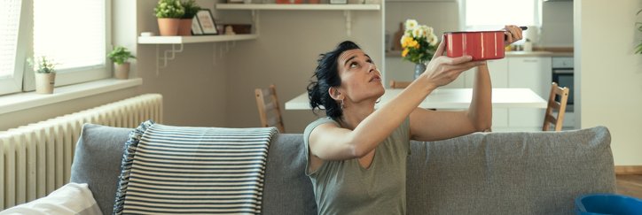A woman sitting on her couch and holding up a pot to catch a leak falling from the ceiling.