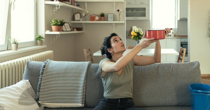 A woman sitting on her couch and holding up a pot to catch a leak falling from the ceiling.