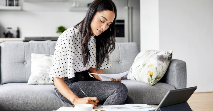 A woman sitting on her couch and looking over paperwork with a tablet open on the coffee table.