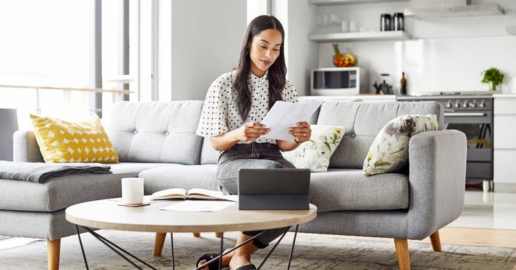 A woman sitting on the couch in her living room reading paperwork in her hand.