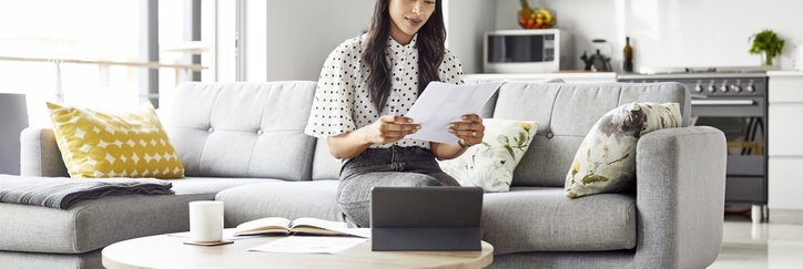 A woman sitting on the couch in her living room reading paperwork in her hand.