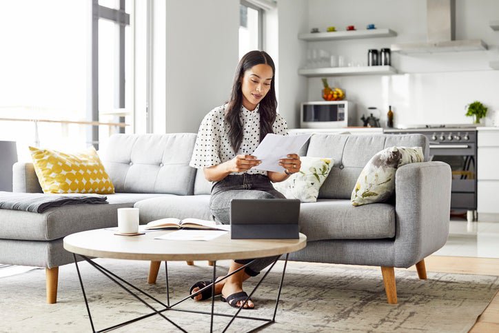 A woman sitting on the couch in her living room reading paperwork in her hand.