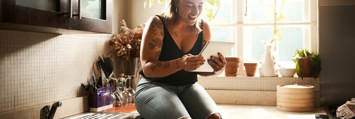 A woman sitting on the kitchen counter in her apartment and smiling at a tablet she's holding.