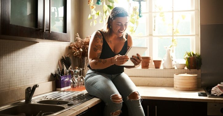 A woman sitting on the kitchen counter in her apartment and smiling at a tablet she's holding.
