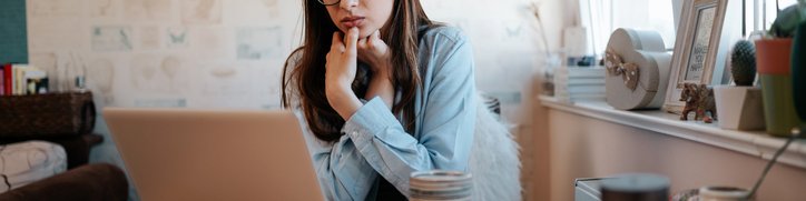 A worried-looking woman looking something up on her laptop while sitting at her kitchen table.