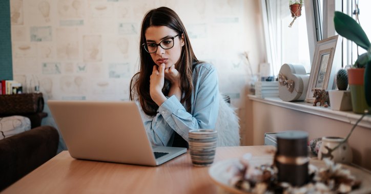 A worried-looking woman looking something up on her laptop while sitting at her kitchen table.