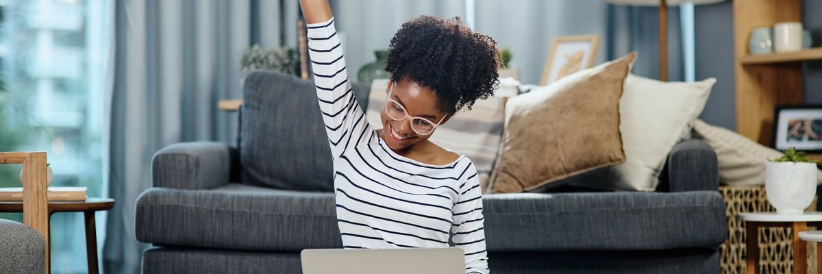 A woman sitting in front of her couch with her laptop and celebrating with her hand in the air.