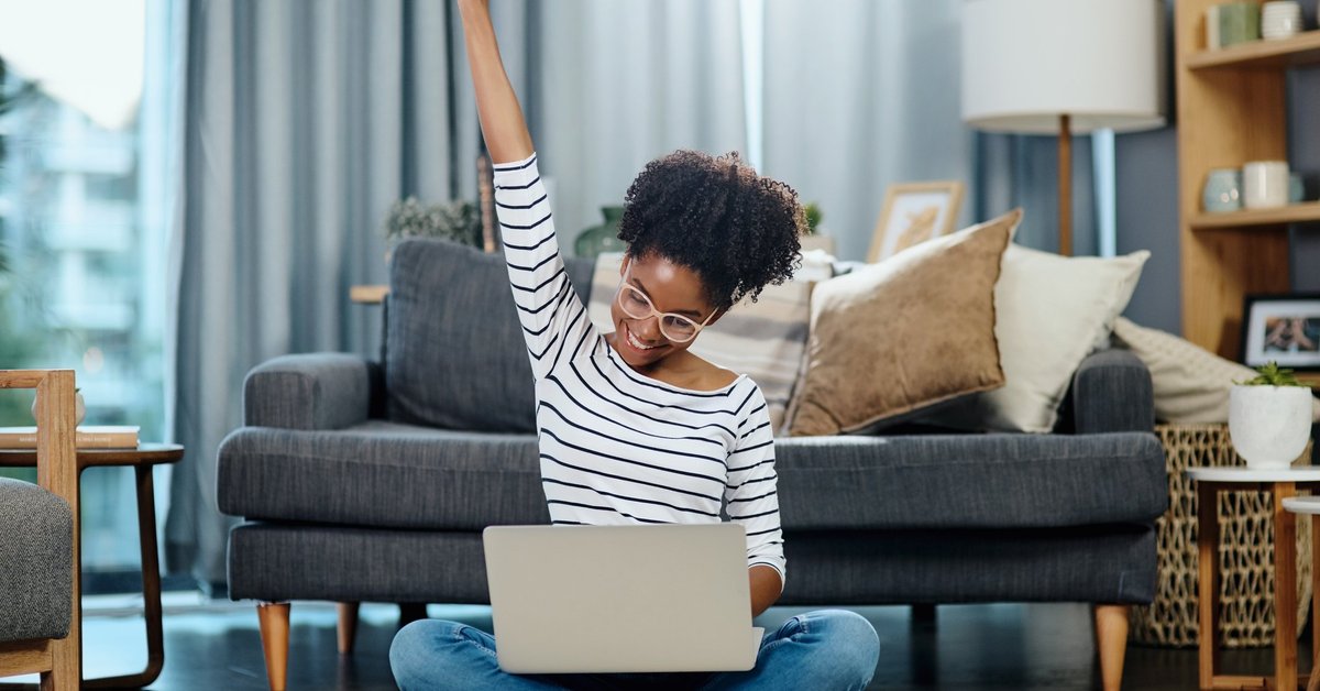 A woman sitting in front of her couch with her laptop and celebrating with her hand in the air.