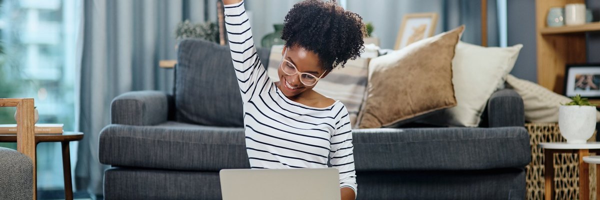 A woman sitting in front of her couch with her laptop and celebrating with her hand in the air.