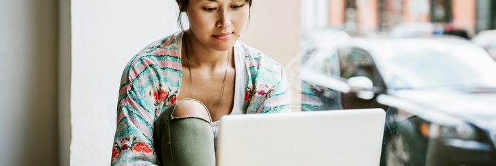 A woman sitting at a table in a cafe window and typing on a laptop.