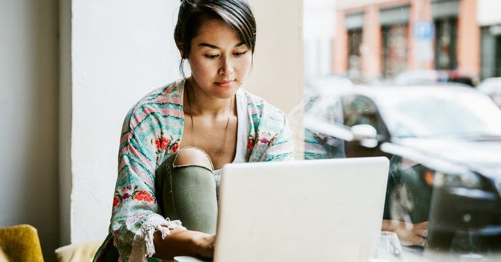 A woman sitting at a table in a cafe window and typing on a laptop.
