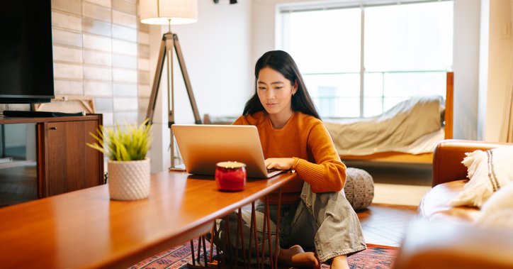 A woman sitting on her living room floor and looking up something on her laptop on the coffee table.