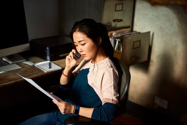 A woman looking at a document while making a phone call from her home office.