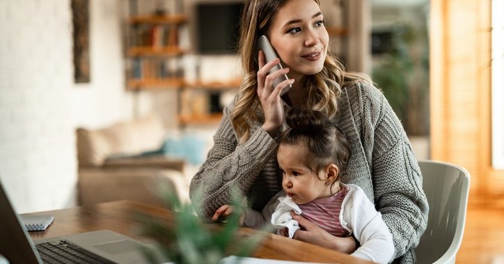 A woman sitting at a desk at home  with her baby in her lap while making a phone call.