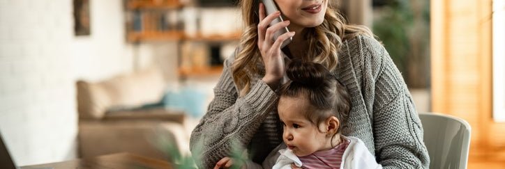 A woman sitting at a desk at home  with her baby in her lap while making a phone call.