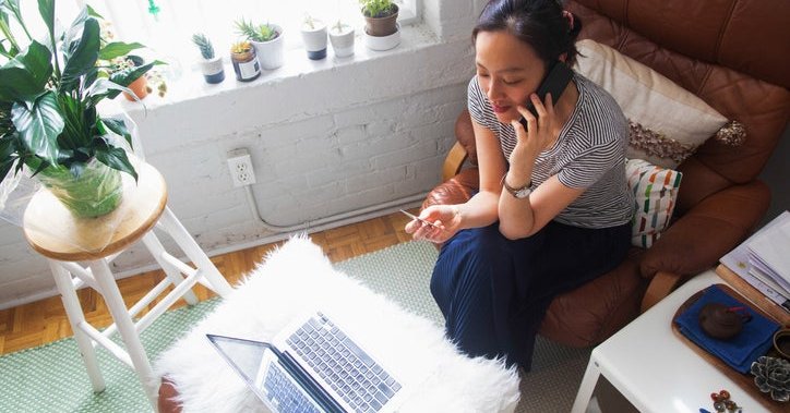 A woman sitting on her couch at home making a phone call while holding a credit card with a laptop open in front of her.