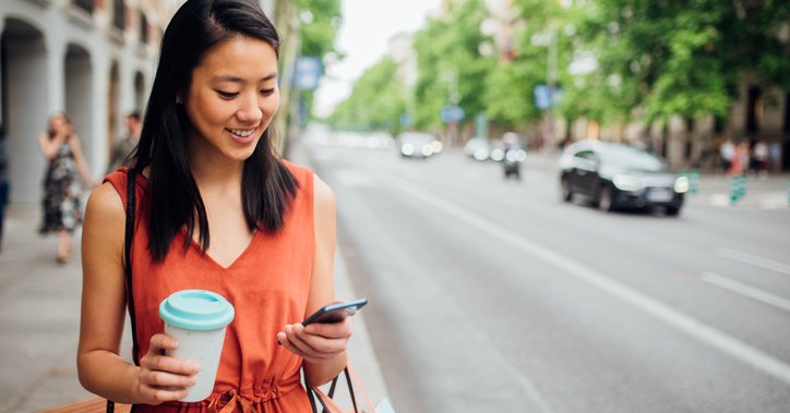 A young woman walking down the street with a coffee in one hand and looking at her phone in her other hand.