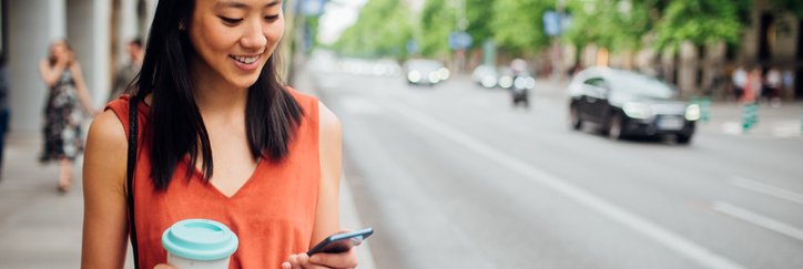 A young woman walking down the street with a coffee in one hand and looking at her phone in her other hand.