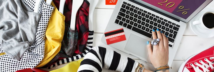 A woman shopping an online sale from her laptop while surrounded by clothes and shoes.