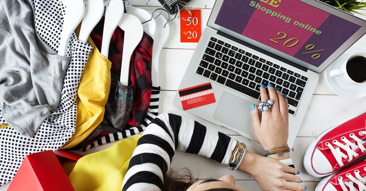 A woman shopping an online sale from her laptop while surrounded by clothes and shoes.