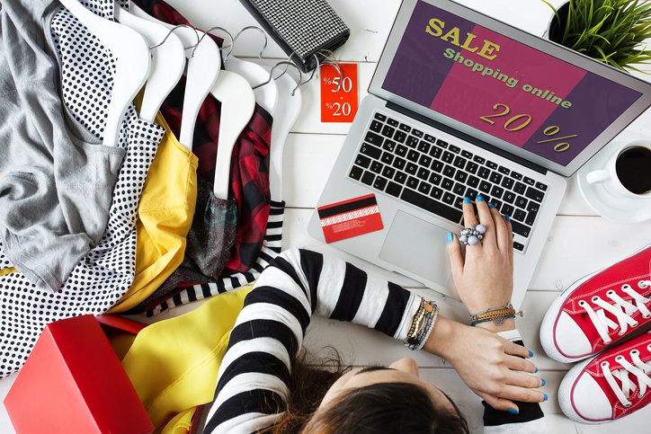 A woman shopping an online sale from her laptop while surrounded by clothes and shoes.