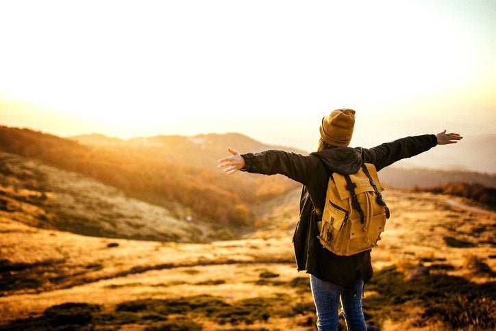A woman in a backpack standing on an open hillside and opening her arms toward the sun in front of her.