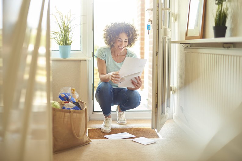 A woman smiling while opening mail in her sunny entryway.