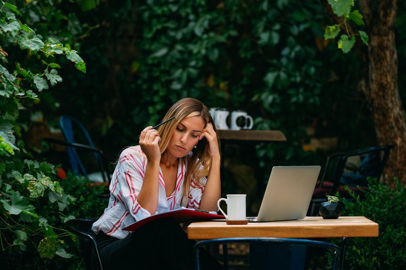 A woman sitting at an outdoor cafe with a laptop and notebook open.
