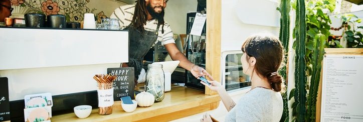 A woman handing her credit card through the window of a food truck to the owner standing inside wearing an apron.